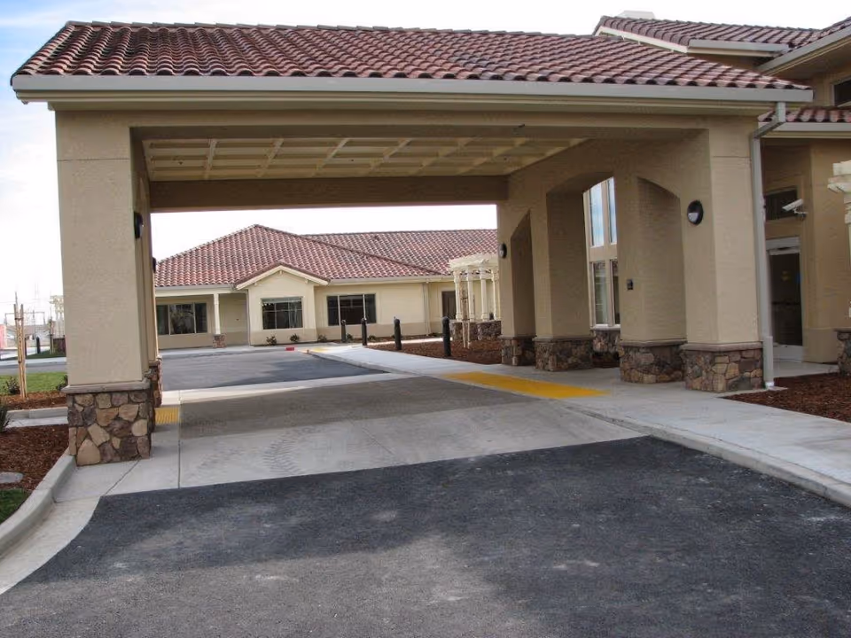 Covered driveway entrance to a building with beige walls, stone accents on the pillars, and red tiled roof. The building appears to be part of a senior living facility with a paved driveway and sidewalk.