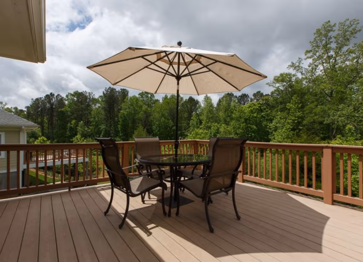 Wooden deck with a round glass table, four chairs and a large patio umbrella overlooking a wooded area.