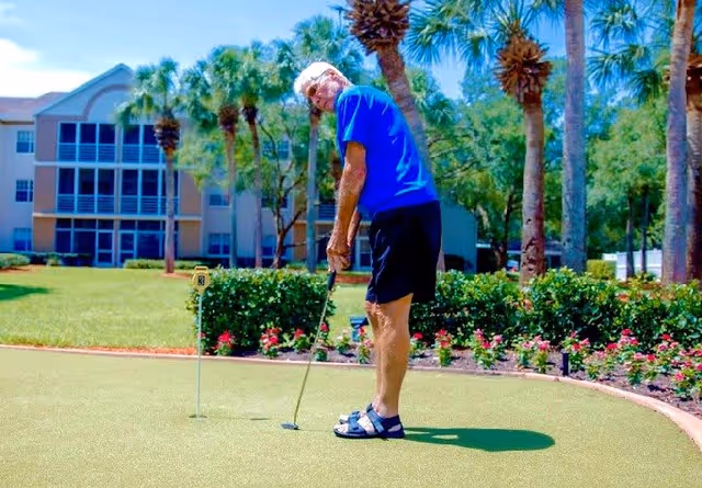 An elderly man wearing a blue shirt, black shorts, and sandals is playing mini golf on a putting green surrounded by palm trees and flowering bushes, with a multi-story building in the background.