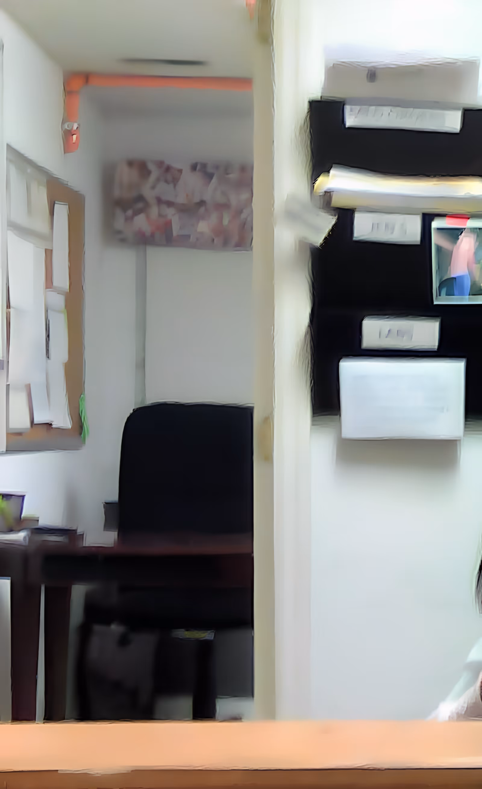 An interior office space with a black office chair, a wooden desk, a bulletin board with papers pinned on the left wall, and a black wall organizer with various papers and a small photo on the right wall.