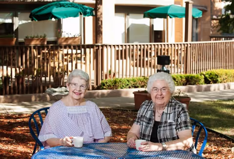 Two elderly women sitting at a blue metal outdoor table with a green umbrella, smiling and holding white cups. They are in a garden area with a wooden fence and plants in the background.