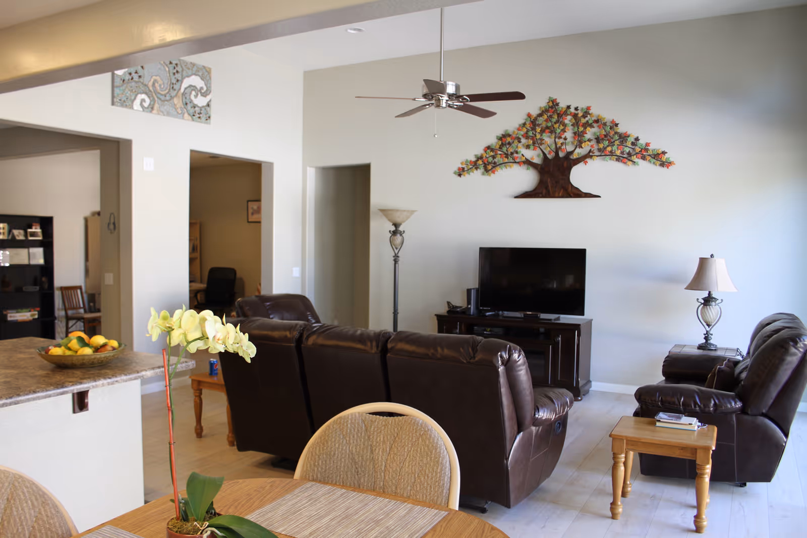 Open-plan living room with brown leather recliners facing a TV under a metal tree wall art, with a dining table and kitchen counter in the foreground.
