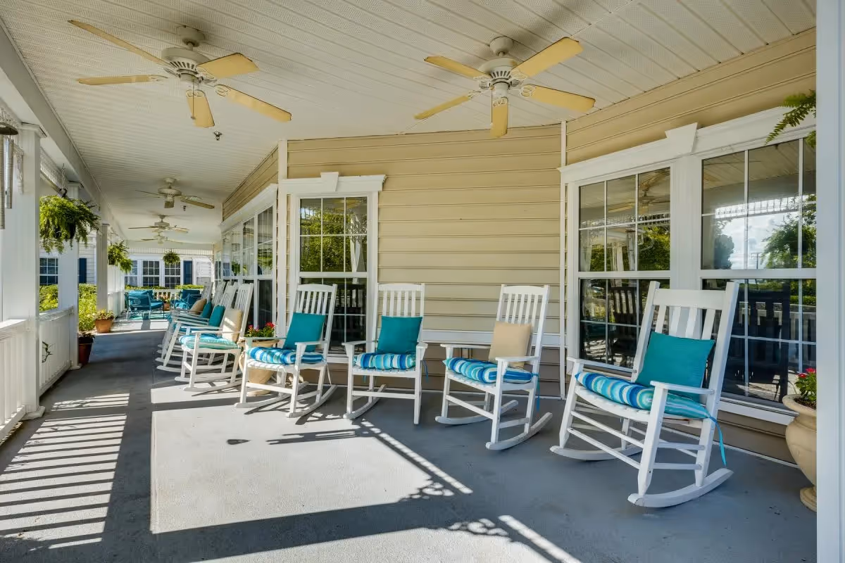 A covered porch area with several white rocking chairs, each with blue and beige cushions, lined up against the beige siding of a building. Ceiling fans are mounted on the white paneled ceiling, and hanging plants are visible along the porch. Large windows reflect the outdoor greenery and sky.