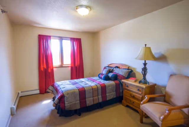 A small bedroom with a single bed covered in a red, black, and beige patterned quilt. The bed has multiple pillows and is positioned against a white wall. Next to the bed is a wooden nightstand with a lamp and a book on it. A beige armchair is placed beside the nightstand. The room has a window with red curtains letting in natural light.