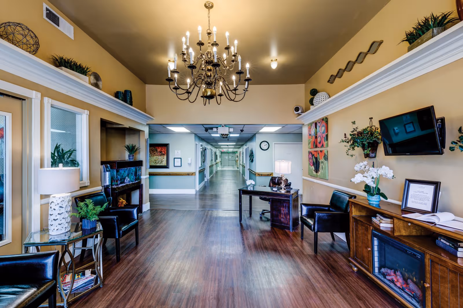 A well-lit interior hallway of a senior living facility with wooden flooring and beige walls. The foreground features a seating area with black leather chairs, a glass side table with a lamp and plant, a wooden cabinet with a fireplace, and a mounted flat-screen TV. A chandelier hangs from the ceiling. The hallway extends into the background with doors and framed artwork along the walls.