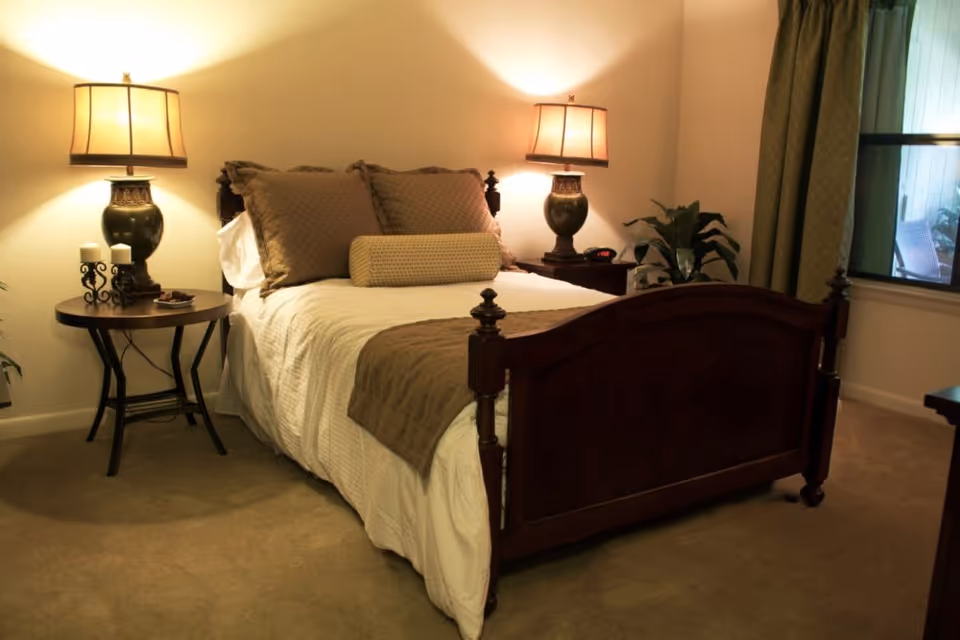 Well-lit bedroom with a wooden bed, decorative pillows, bedside tables with lamps, and a window with green curtains.
