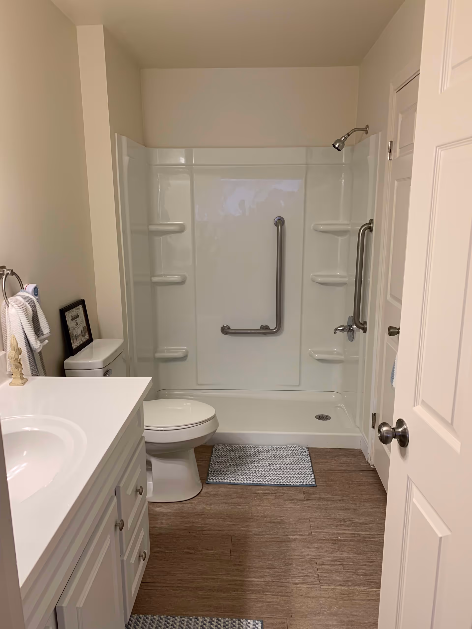A clean bathroom with a white vanity and sink on the left, a toilet next to it, and a walk-in shower with grab bars and built-in shelves at the back. The floor has wood-like tiles with two small rugs, and the walls are painted a light beige color.