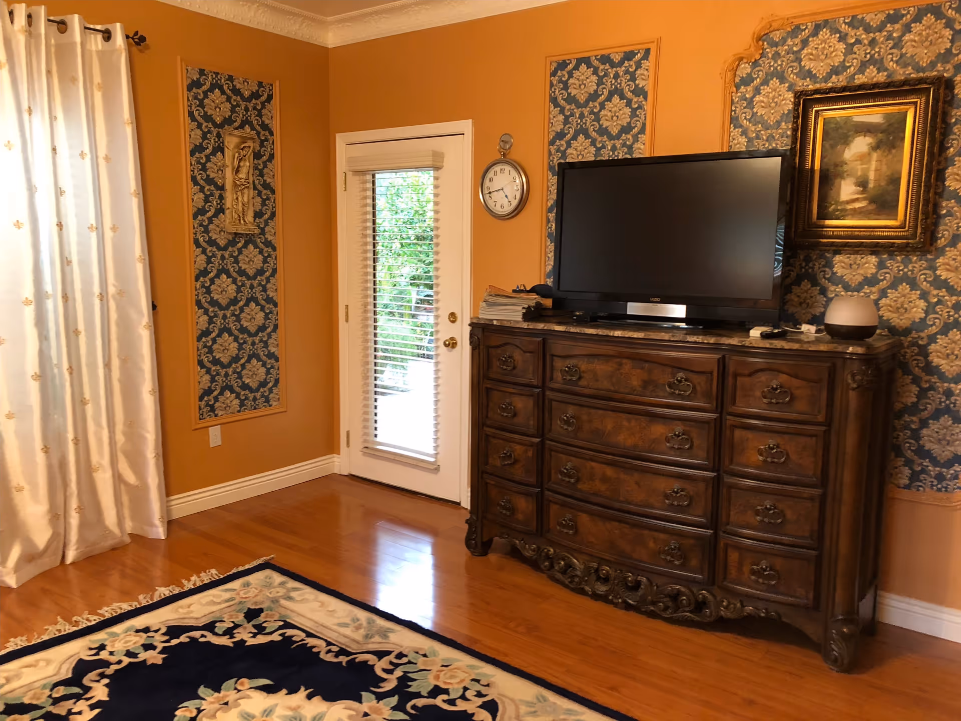 A cozy living room with warm orange walls and blue patterned wallpaper panels. There is a wooden dresser with ornate carvings holding a flat-screen TV, a clock on the wall, a framed painting, and a small smart speaker. A glass door with blinds leads outside, and a decorative rug covers part of the wooden floor. White curtains with a subtle pattern hang on the left side.
