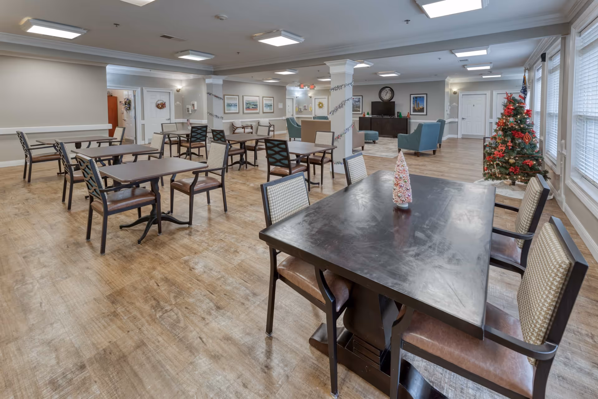 A spacious dining and common area in a senior living facility with multiple tables and chairs arranged on a wood floor. The room is decorated for the holidays with a Christmas tree and a small decorative Christmas tree on one table. The walls are adorned with framed pictures, and there is a television with a clock above it in the background. Large windows on the right side let in natural light.