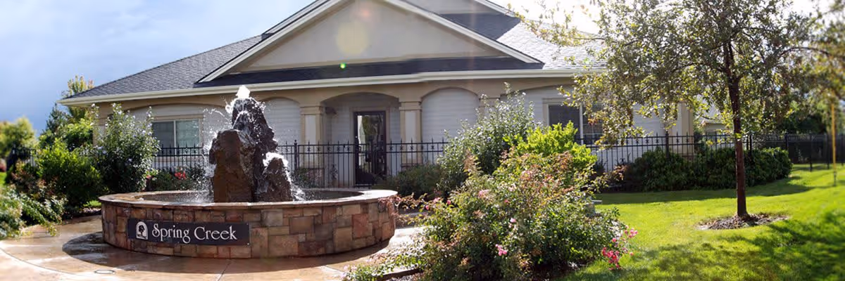 Outdoor view of a building with a stone fountain in front labeled 'Spring Creek'. The area is landscaped with green grass, bushes, and trees under a partly cloudy sky.