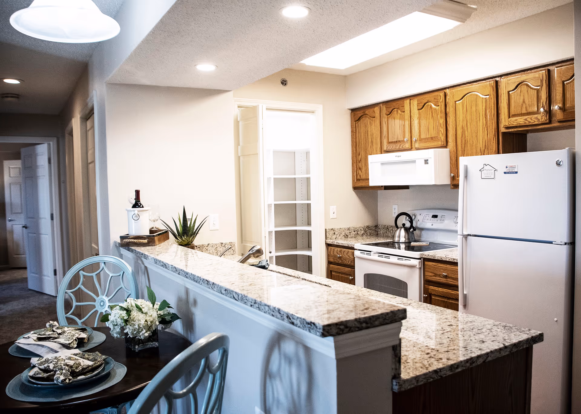 Bright kitchen with a granite countertop bar, wooden cabinets, white appliances, and a small dining area set for two.