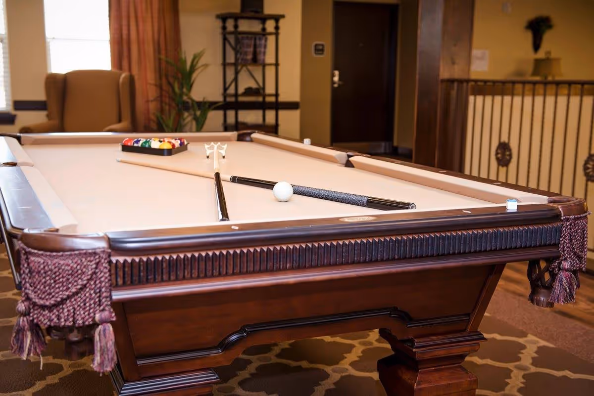 A billiards table with a beige felt surface in a cozy room. The table has a set of pool balls racked at one end, two pool cues crossed near the center, and a white cue ball. In the background, there is a comfortable armchair, a plant, a shelving unit, and a railing with decorative elements. The room has warm lighting and a patterned carpet.