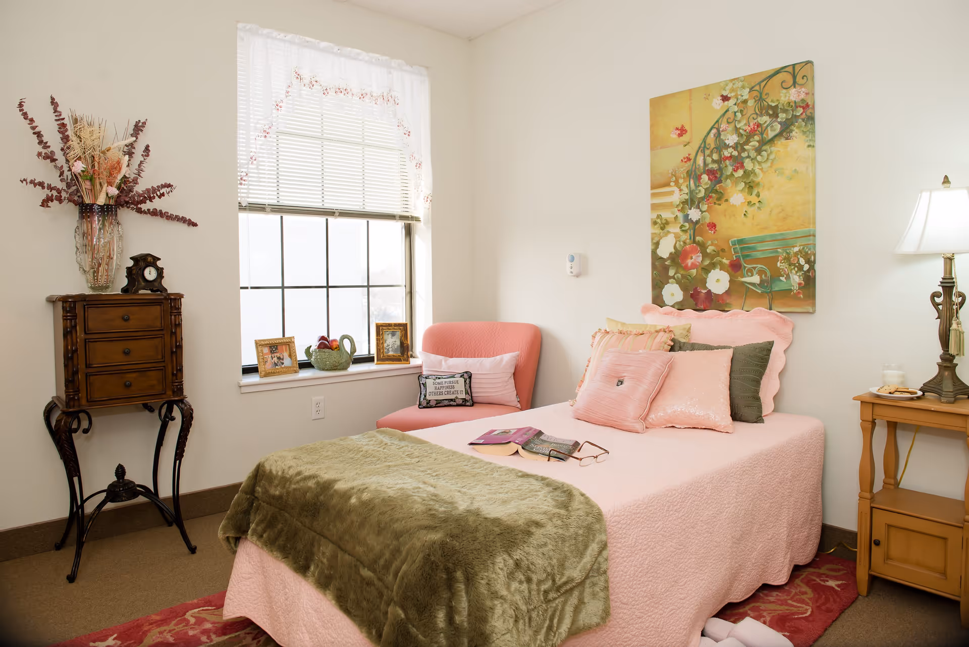A cozy bedroom with a pink bedspread and multiple pillows, a green throw blanket, an open book and glasses on the bed. There is a pink upholstered chair next to a window with white blinds and lace curtains. A wooden side table with a lamp and a small clock on a decorative stand are also visible. A floral painting hangs on the wall above the bed.