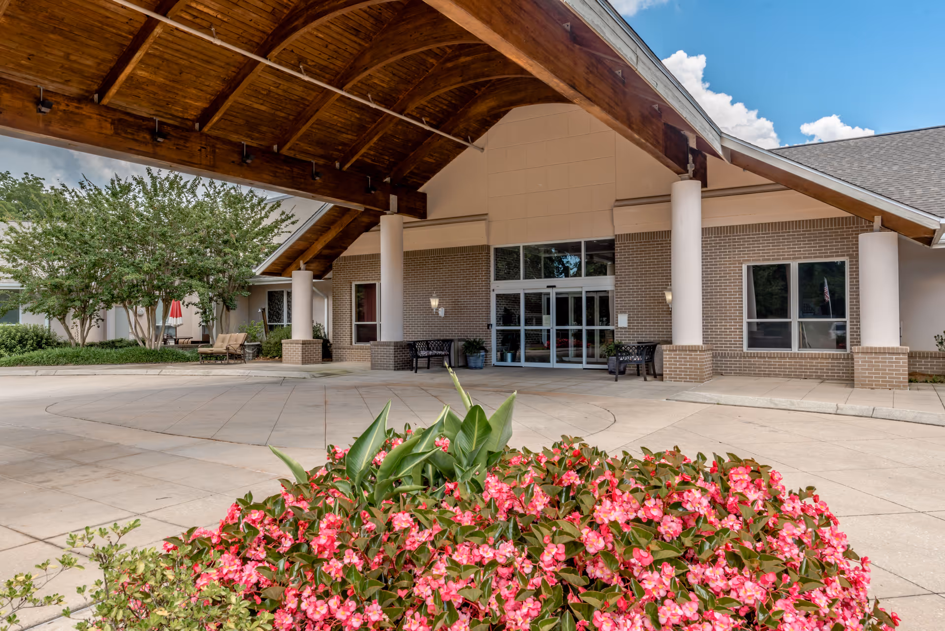 Entrance of a senior living facility with a covered driveway supported by large white columns. There are benches near the entrance, a flower bed with pink flowers in the foreground, and trees and shrubs on the left side. The building has a brick facade and large glass doors under a wooden roof structure.
