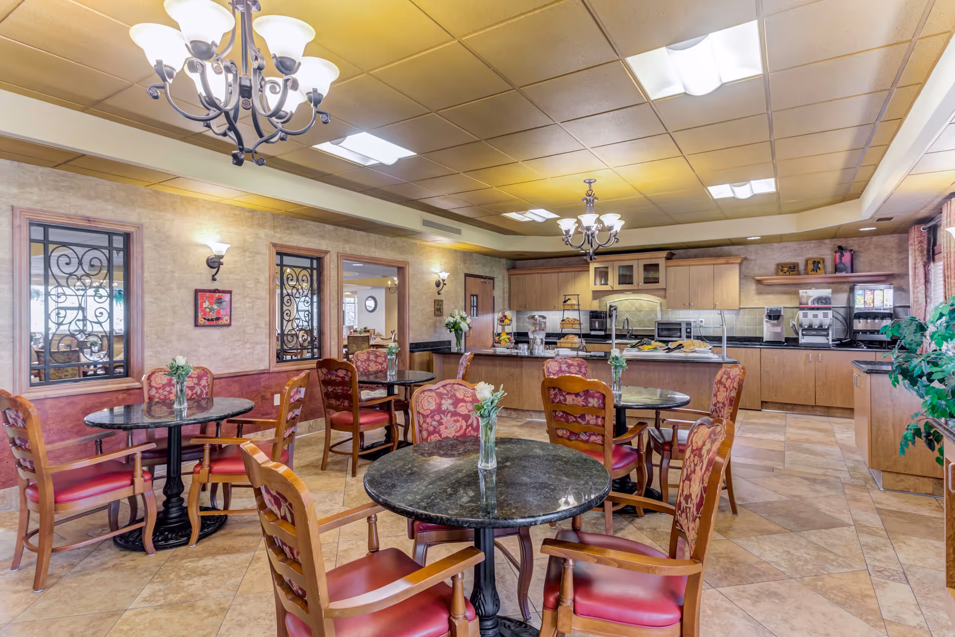 A cozy dining area in Chandler Place featuring round tables with floral upholstered chairs. Each table has a small vase with flowers. The room has warm lighting with chandeliers and wall sconces, and a kitchen area with wooden cabinets and various appliances is visible in the background.