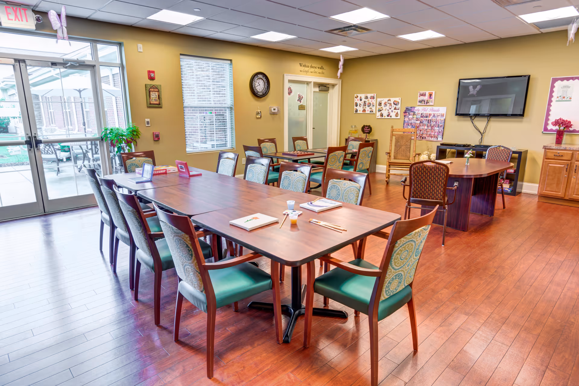 A bright and spacious common room with wooden floors and multiple tables surrounded by chairs. The room has a large window and glass doors leading to an outdoor patio area. There is a wall-mounted TV, a clock, and various decorations on the walls. The room appears clean and inviting, suitable for group activities or dining.
