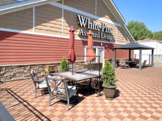 Outdoor patio area at White Pine Advanced Assisted Living and Memory Care - Fridley with tables, chairs, umbrellas, potted plants, and a gazebo against the building exterior.