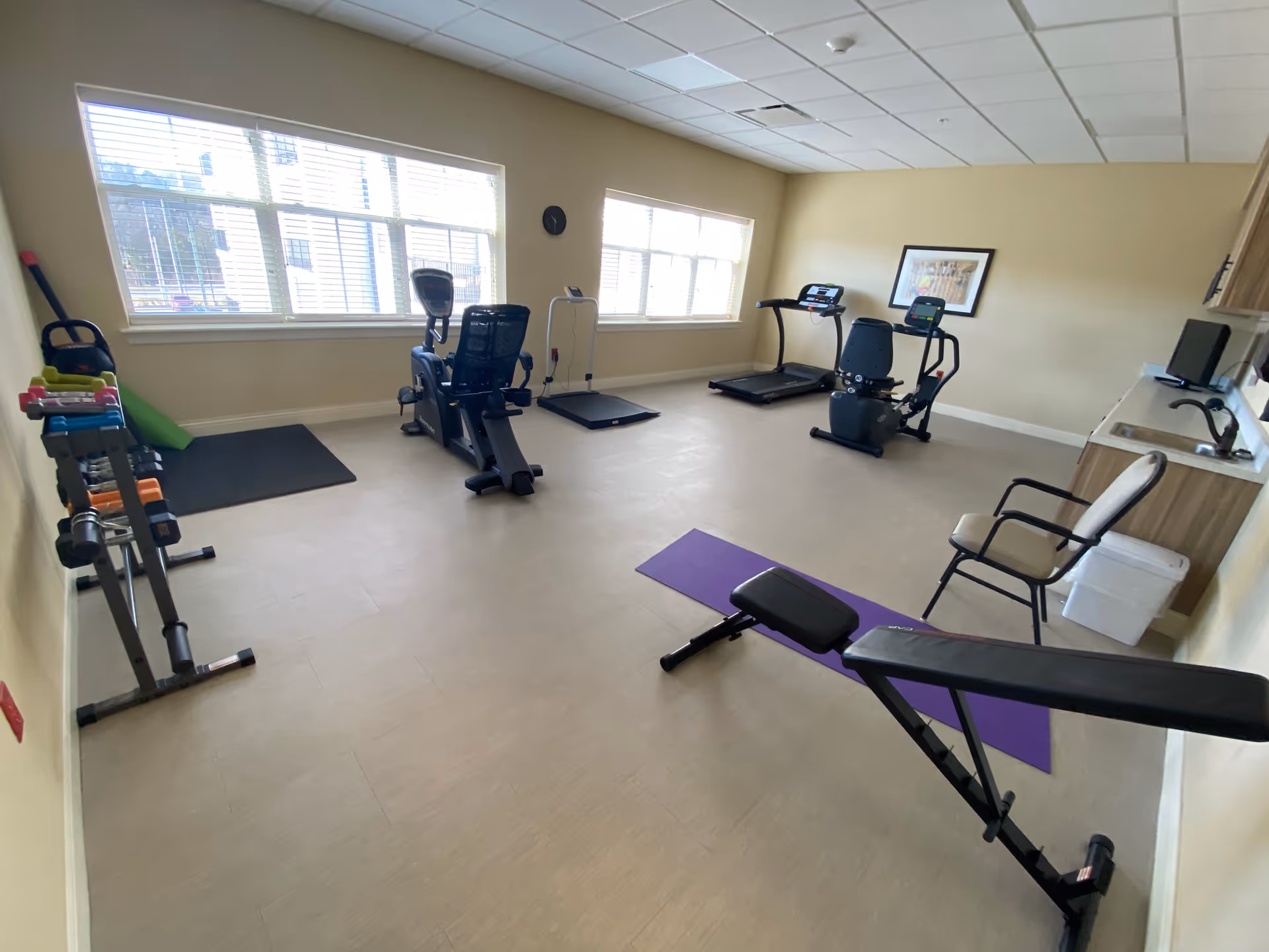 Bright fitness room with treadmills, a recumbent bike, weight bench, dumbbells and a yoga mat under large windows.