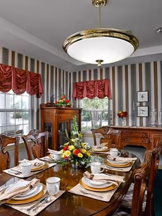 A formal dining room with a polished wooden table set for six people, featuring white plates, cups, and napkins on placemats. The room has striped wallpaper, red valance curtains on the windows, a wooden sideboard with decorative items, and a large round ceiling light fixture.