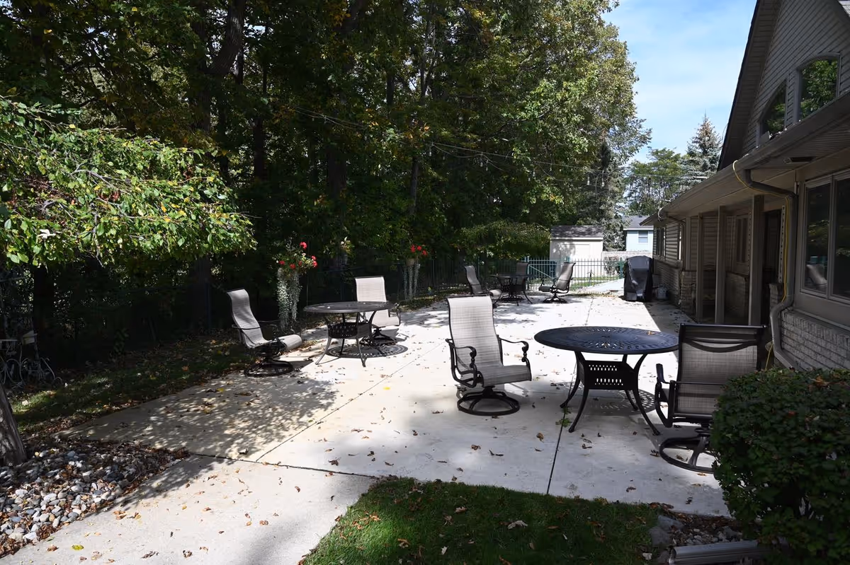 Outdoor patio area at Leisure Manor Residence for Seniors with several round metal tables and cushioned swivel chairs arranged on a concrete surface. The patio is adjacent to a building and surrounded by trees and greenery, with some potted plants and a small shed visible in the background under a clear sky.