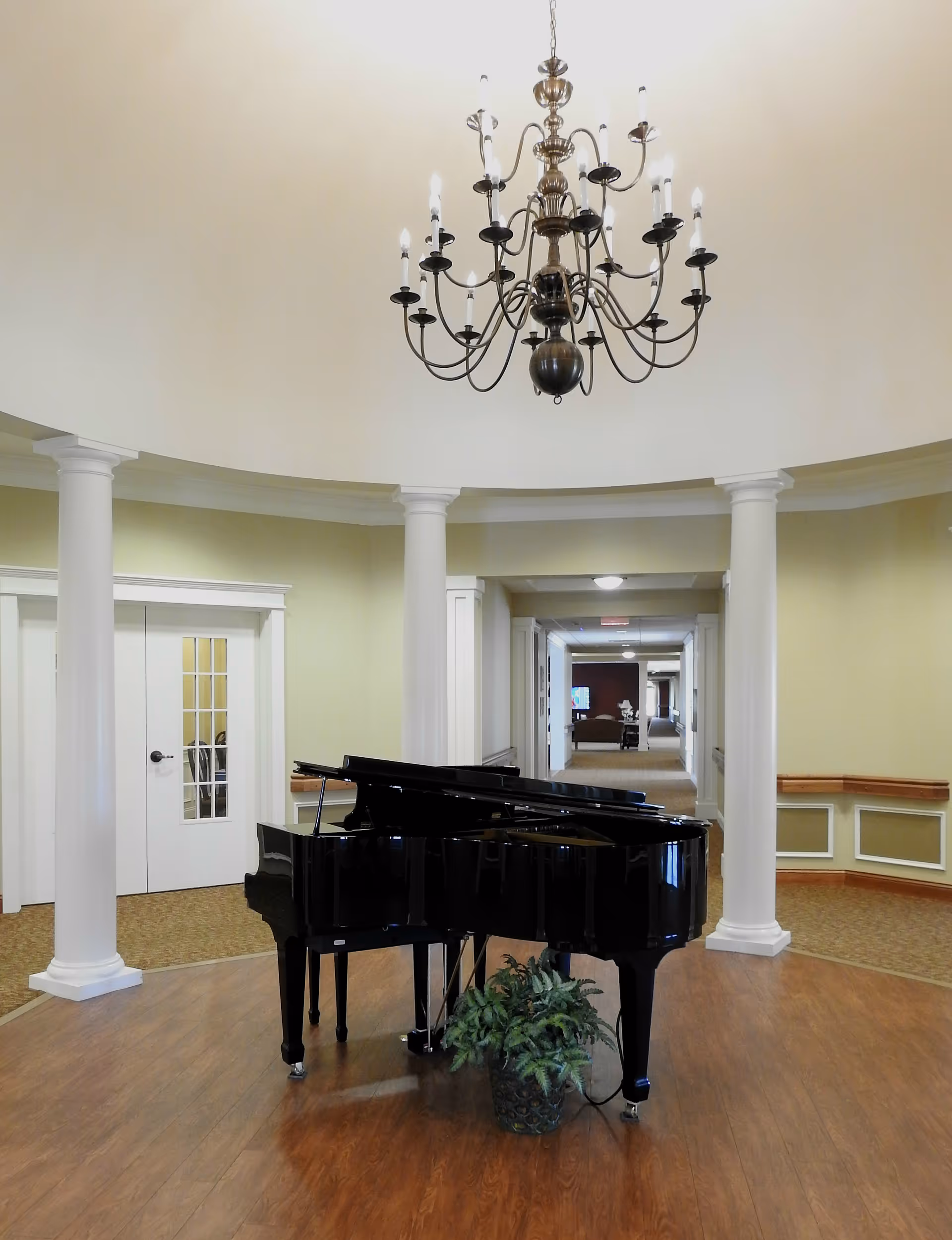 A black grand piano beneath a large chandelier in a circular lobby with white columns and a central hallway.