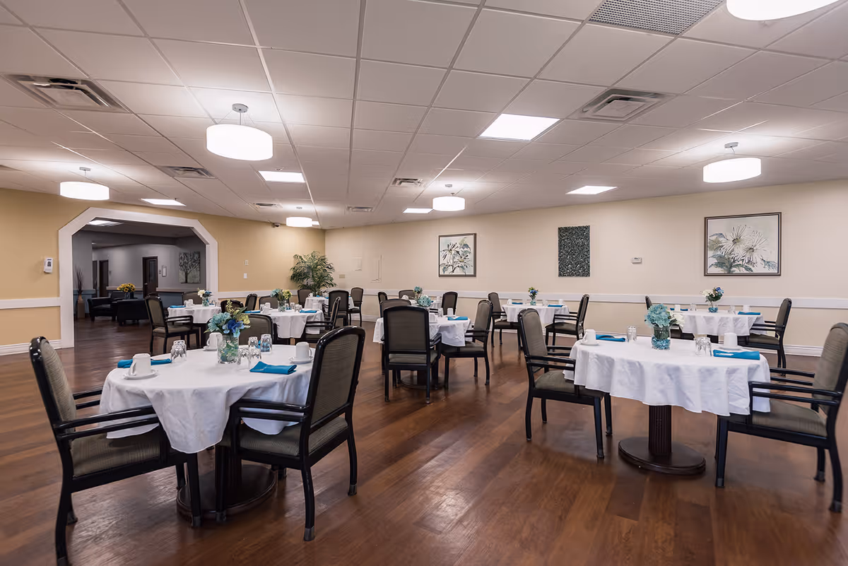 Spacious dining room with round tables covered in white tablecloths, place settings, and chairs on wood flooring.
