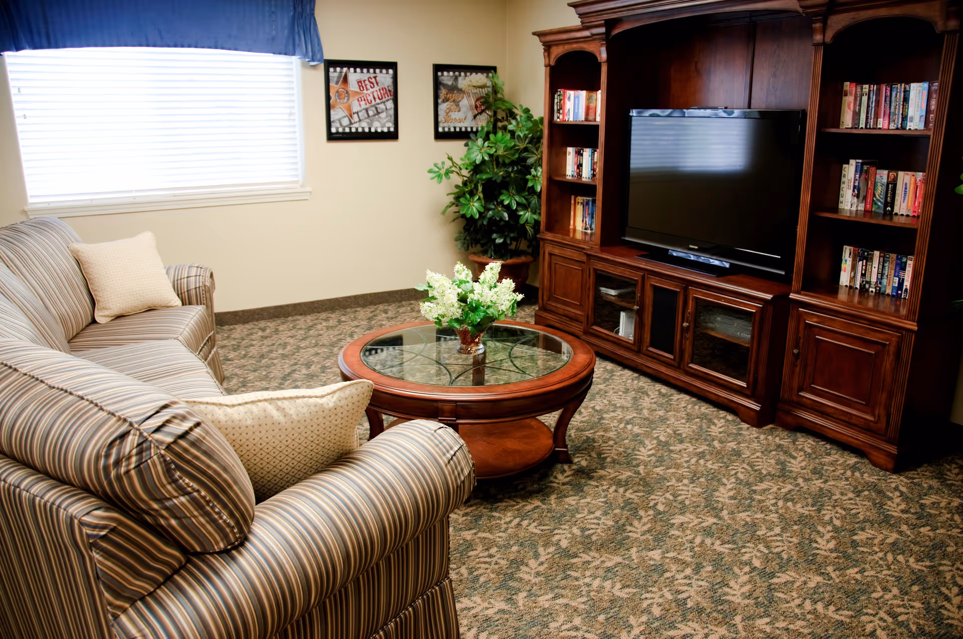 A cozy living room with a striped sofa and armchair, a round glass-top wooden coffee table with a flower arrangement, a large wooden entertainment center with a flat-screen TV and bookshelves, a window with blinds and a blue valance, two framed pictures on the wall, and a potted plant in the corner.