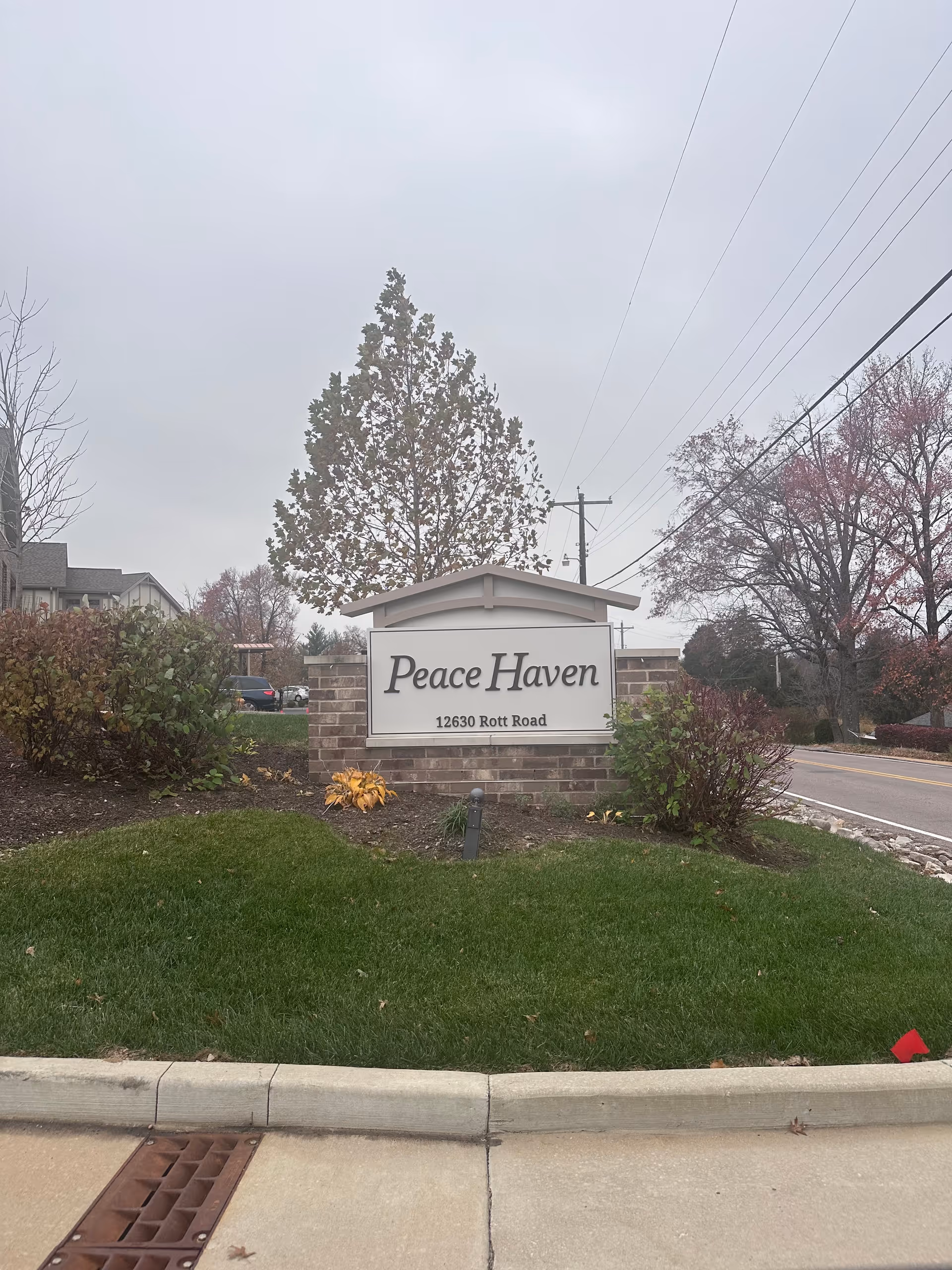 Outdoor view of a brick sign for Peace Haven located at 12630 Rott Road, surrounded by green grass, bushes, and trees with a cloudy sky in the background.