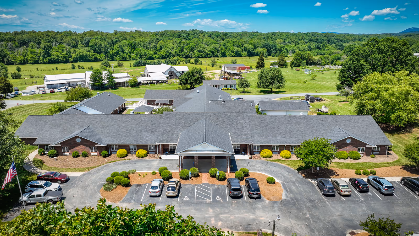 Aerial view of Walnut Ridge Assisted Living facility showing a single-story brick building with a large parking lot in front. The building has a covered entrance and is surrounded by landscaped bushes and trees. In the background, there are green fields, trees, and a few farm buildings under a blue sky with scattered clouds.