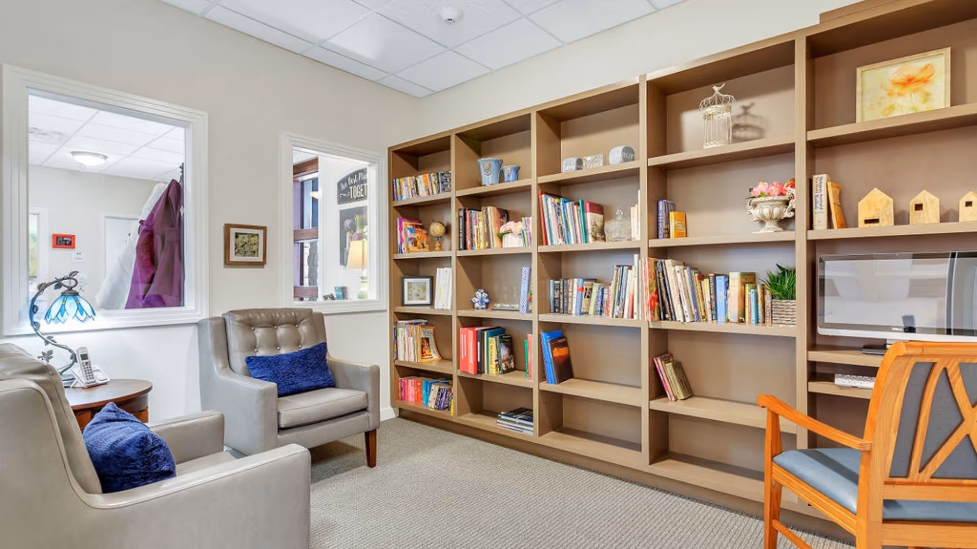 A cozy reading room with two gray armchairs each with a blue cushion, a wooden bookshelf filled with books and decorative items, a wooden chair with blue upholstery, and a small table with a decorative lamp and a phone. The room has beige walls and carpeted floor.