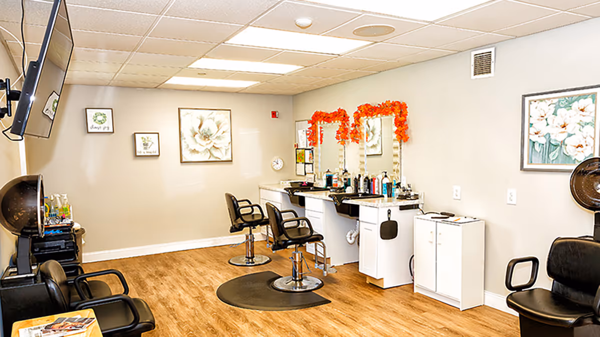 Interior of a salon area in a senior living facility with two black styling chairs in front of mirrors decorated with orange garlands. The room has light-colored walls with floral artwork, a wooden floor, and salon equipment including hair dryers and various hair care products.