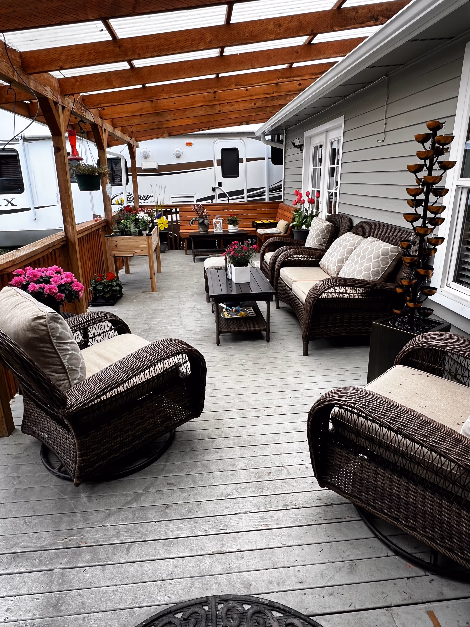 Covered wooden patio with wicker chairs, a coffee table and potted flowers next to a house and a parked RV.