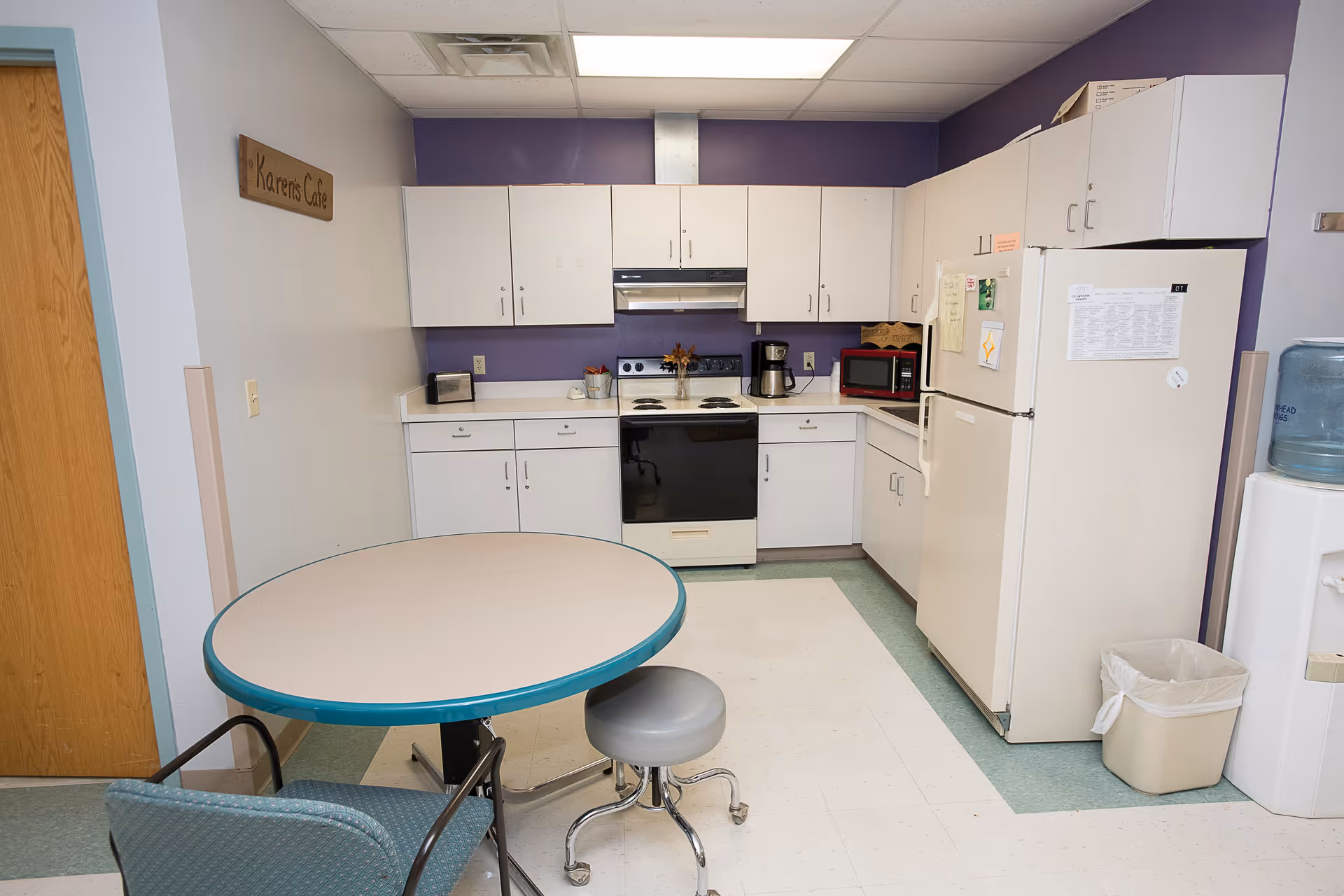 A small kitchen area with white cabinets, a stove, a refrigerator, a microwave, a coffee maker, and a water cooler. There is a round table with a teal edge and two chairs, one with armrests and one stool. A wooden sign on the wall reads 'Karens Cafe'. The walls are painted purple and white.