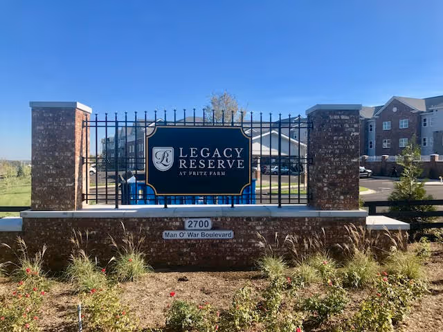 Entrance sign for Legacy Reserve at Fritz Farm, mounted on a brick and metal fence structure with landscaping in front and residential buildings visible in the background under a clear blue sky.