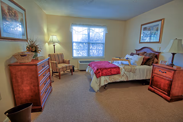 A cozy bedroom in a senior living facility with a bed covered in beige and red bedding, a wooden nightstand with a lamp, a wooden dresser with a basket and plant, a striped armchair, a window with blinds and a valance, and framed artwork on the walls.