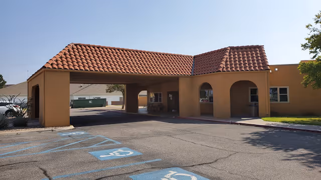 Exterior view of a building entrance with a covered driveway featuring a red tile roof. The building has a tan stucco finish and arched openings. There are several handicap parking spaces marked in blue in the foreground, and a tree is visible on the right side.