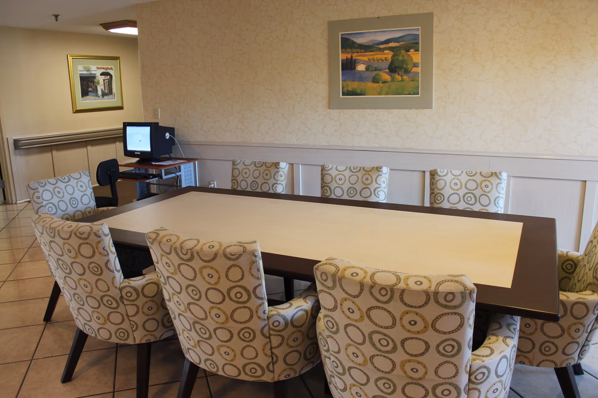 A conference or meeting room with a rectangular table surrounded by eight cushioned chairs with circular patterned upholstery. The room has beige walls with a framed landscape painting and a framed picture on adjacent walls. A computer workstation with a monitor and chair is visible in the corner.