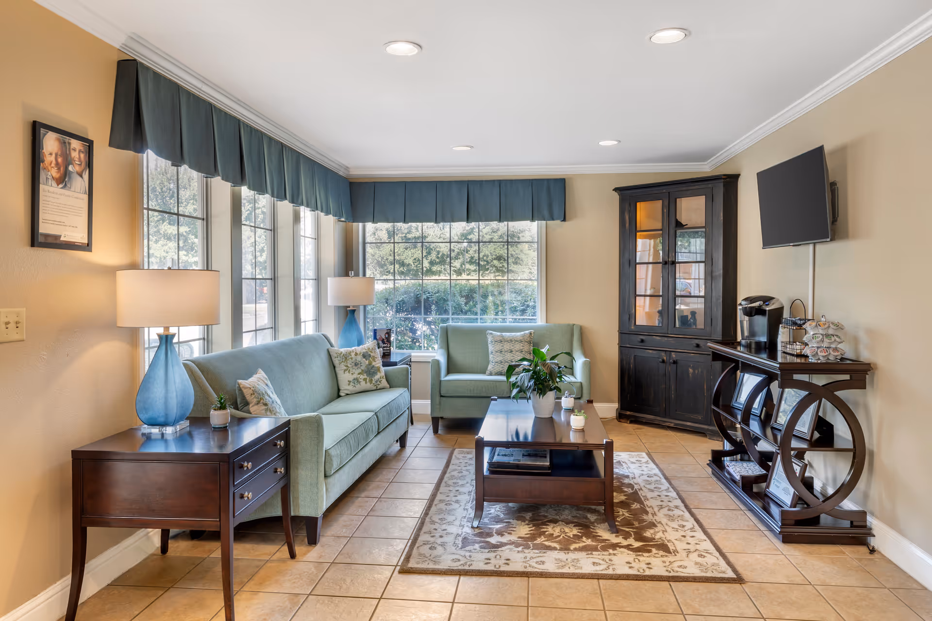 A cozy living room with two light green sofas, a wooden coffee table on a patterned rug, two side tables with blue lamps, a dark wooden cabinet, a TV mounted on the wall, and a coffee station with a Keurig machine. Large windows with blue valances let in natural light and show greenery outside.
