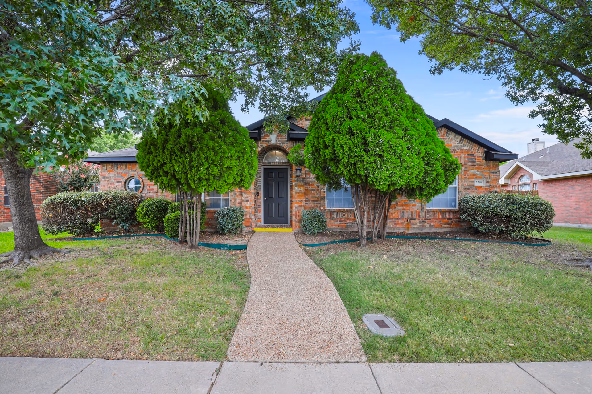 Front exterior view of a single-story brick house with a black front door, two large green trees flanking the entrance, trimmed bushes, and a concrete walkway leading from the sidewalk to the door.