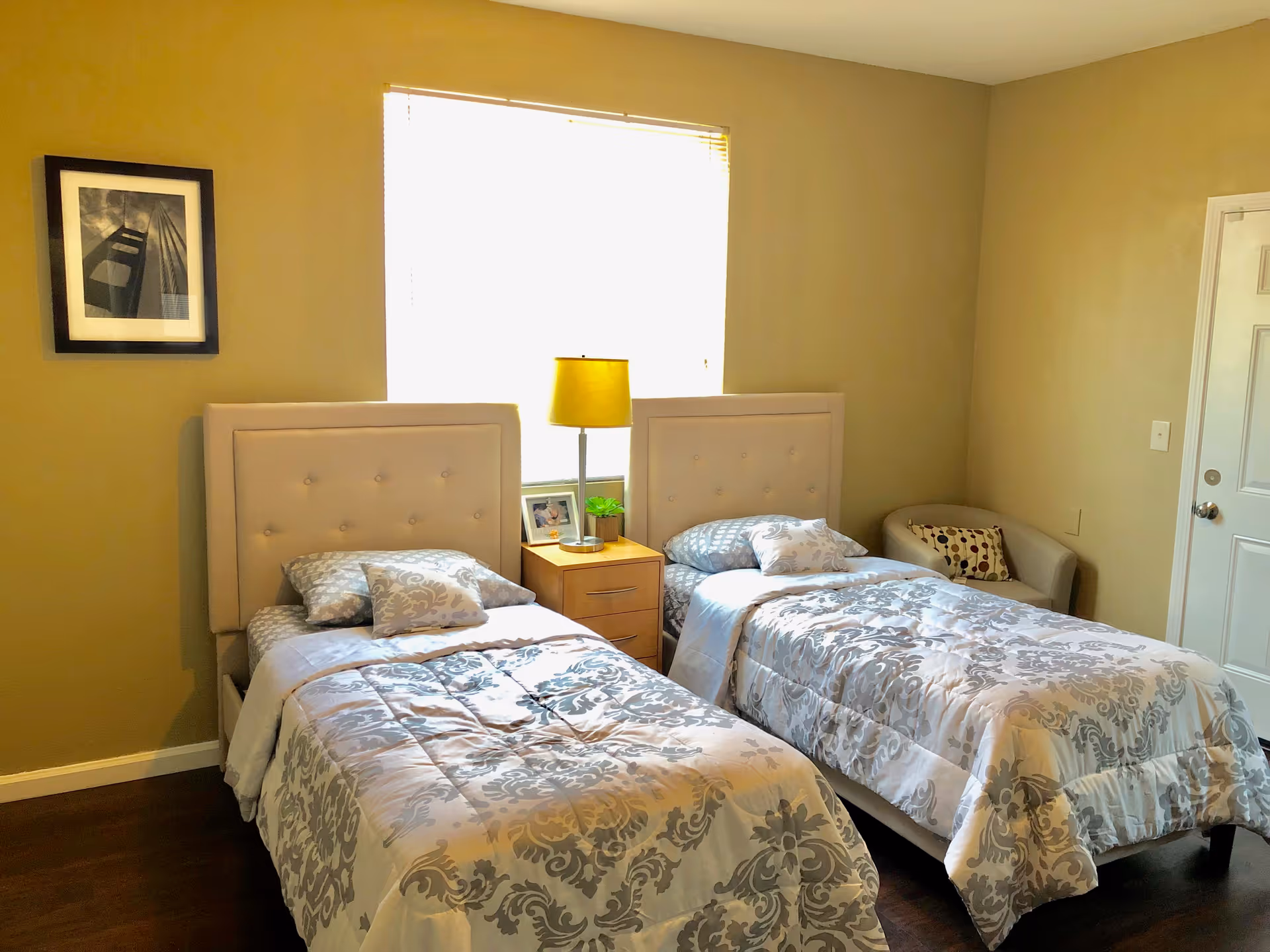 A bedroom with two twin beds featuring light-colored tufted headboards and matching patterned bedding. Between the beds is a wooden nightstand with a yellow lamp, a small plant, and a framed photo. A window with blinds is centered behind the nightstand. On the right side of the room is a small beige armchair with a decorative pillow. The walls are painted a warm beige color, and there is a framed black and white photograph on the left wall.