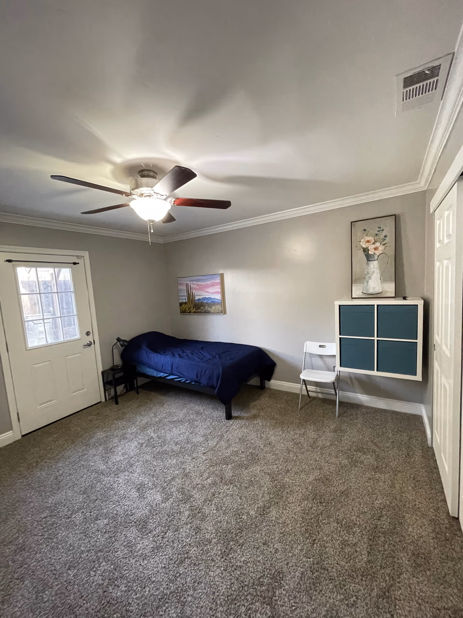 A small bedroom with beige carpet and light gray walls. There is a single bed with a dark blue blanket against one wall, a small black nightstand with a lamp next to it, and a white folding chair next to a wall-mounted storage unit with four blue compartments. Two framed pictures hang on the walls, one above the bed showing a desert scene with cacti and mountains, and another above the storage unit depicting flowers in a pitcher. A ceiling fan with a light is on the ceiling, and a door with a window leads outside.