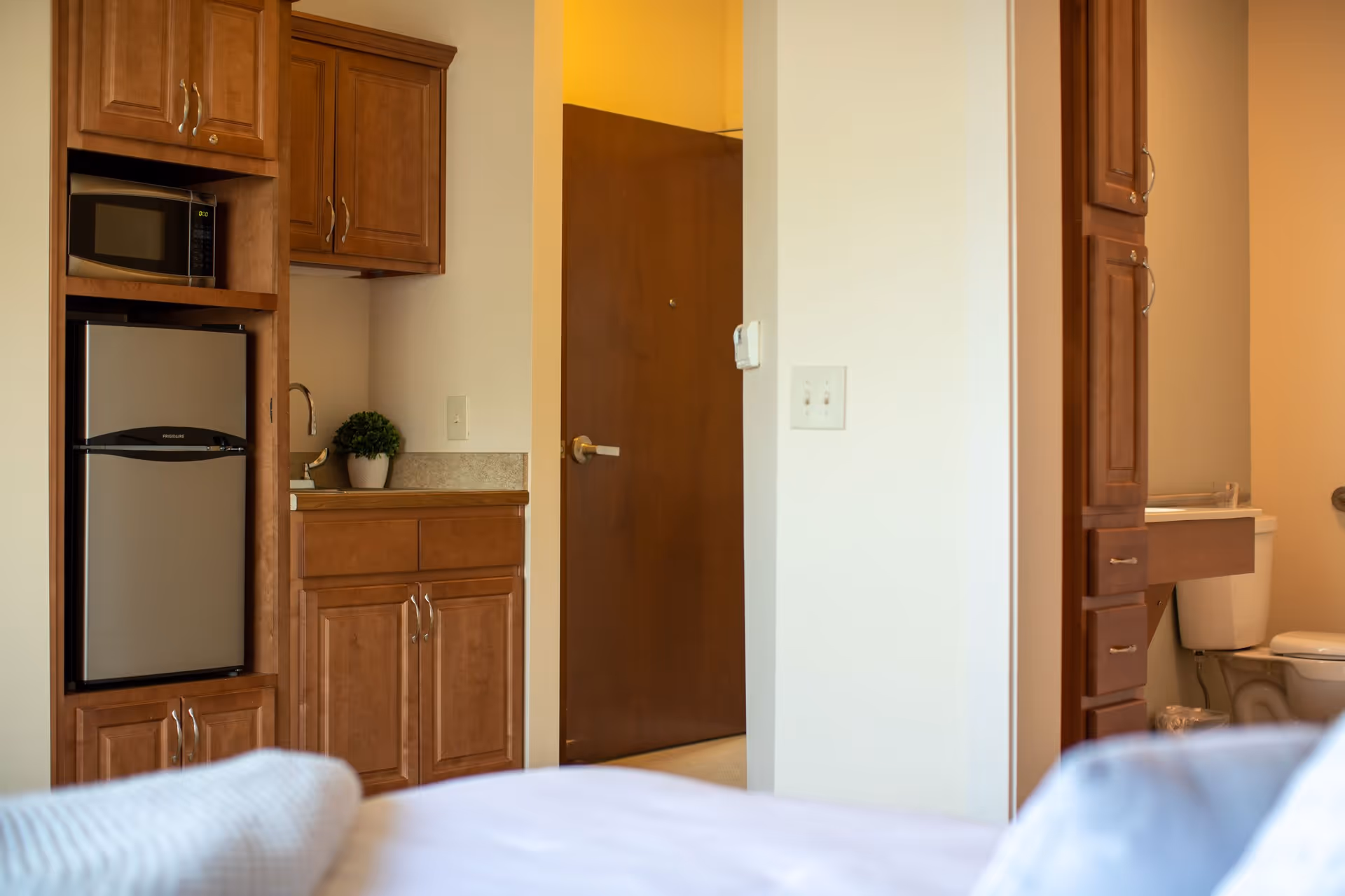 Studio-style apartment interior showing a small kitchenette with microwave, mini fridge and cabinets, a doorway and a bathroom with a toilet, with part of a bed in the foreground.