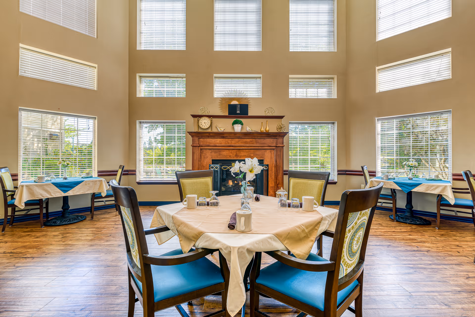 A bright dining room with multiple tables covered in beige tablecloths and blue table runners. Each table is set with cups, glasses, and a vase with white flowers. The room features large windows with white blinds and a wooden fireplace with decorative items on the mantel.