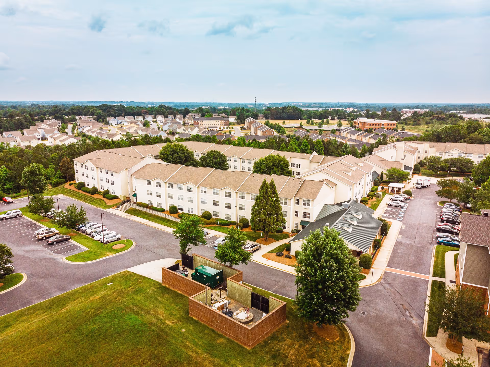 Aerial view of a senior living facility named The Dorchester, showing multiple connected beige buildings with brown roofs, surrounded by parking lots, trees, and green lawns. Residential houses and other buildings are visible in the background under a partly cloudy sky.