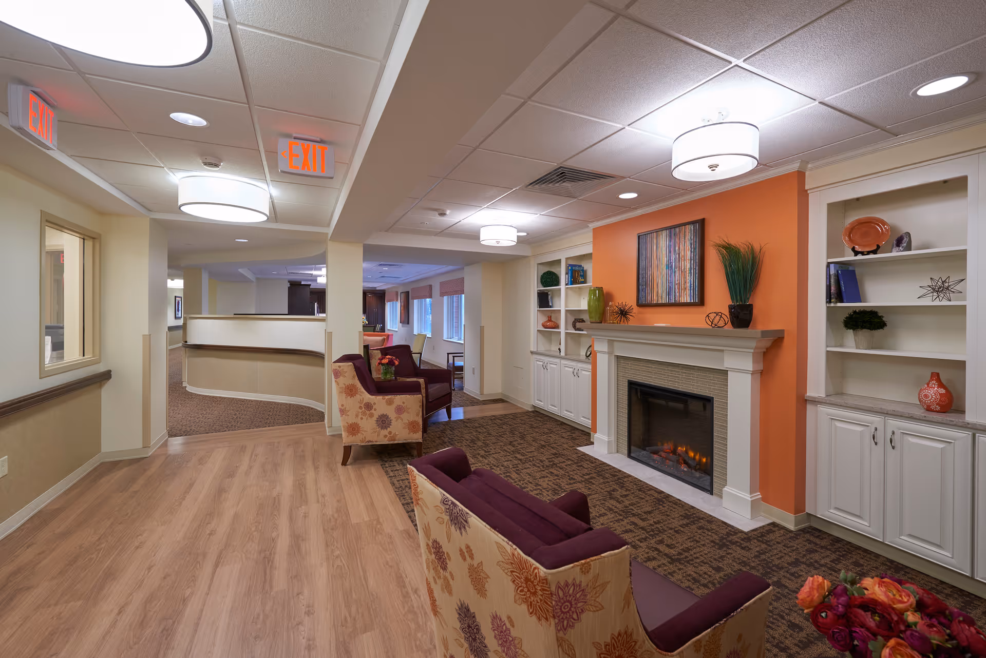 A cozy common area in a senior living facility featuring a fireplace with a bright orange accent wall, built-in white shelves decorated with vases and books, and comfortable armchairs with floral upholstery. The space has wood flooring, carpeted area near the fireplace, ceiling lights, and exit signs visible in the hallway.