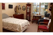 Cozy furnished bedroom with a bed, dresser, wall-mounted TV, display cabinet, chairs, and a window.