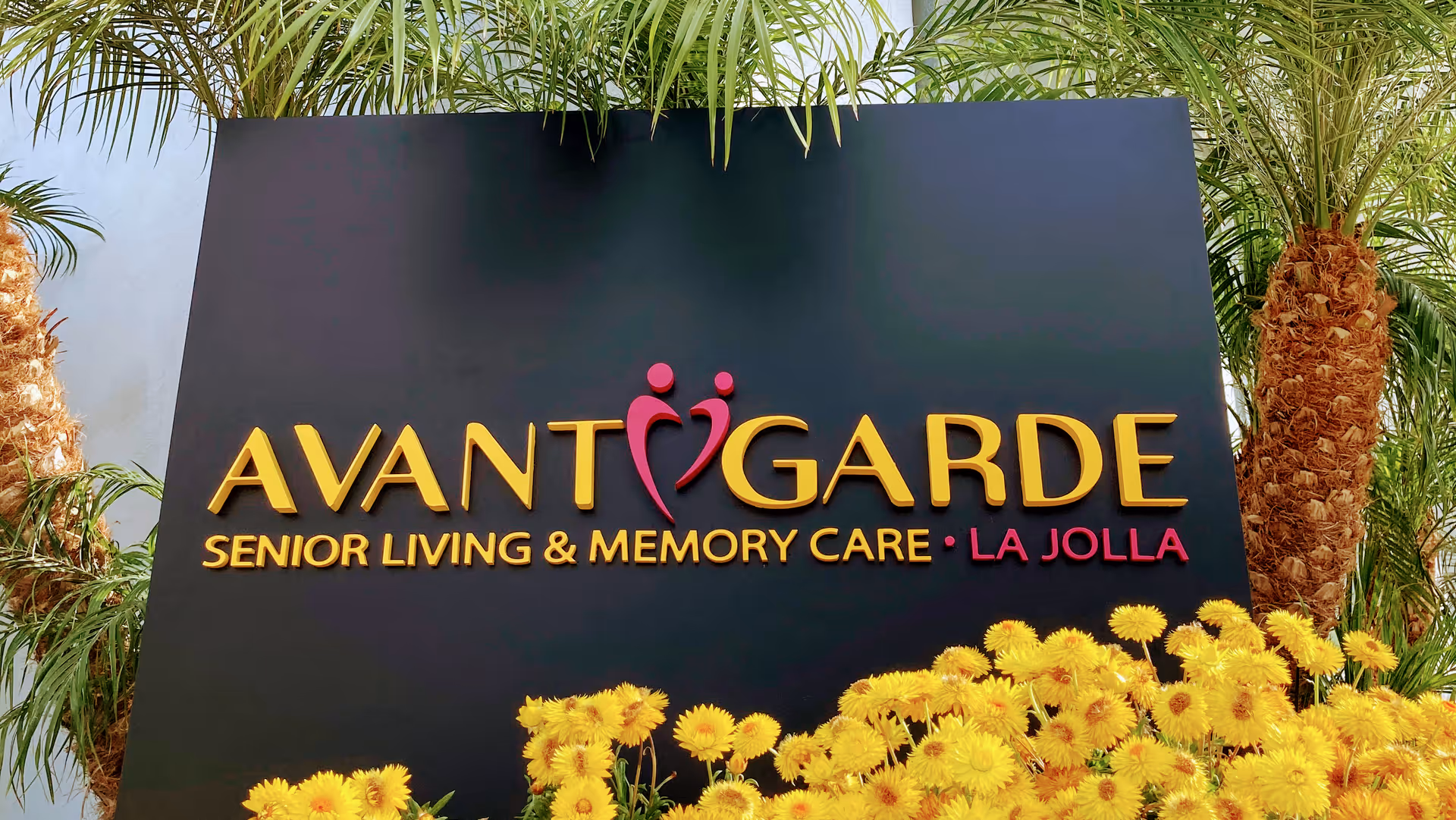 Entrance sign reading Avantgarde Senior Living & Memory Care • La Jolla surrounded by palm trees and yellow flowers.