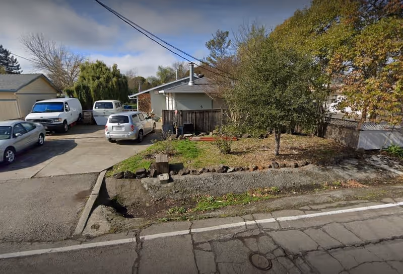 Exterior view of a single-story residential building with a driveway containing several parked vehicles. There is a small front yard with grass, a tree, and some shrubs. The street in front shows cracked pavement and a white line marking the edge of the road.