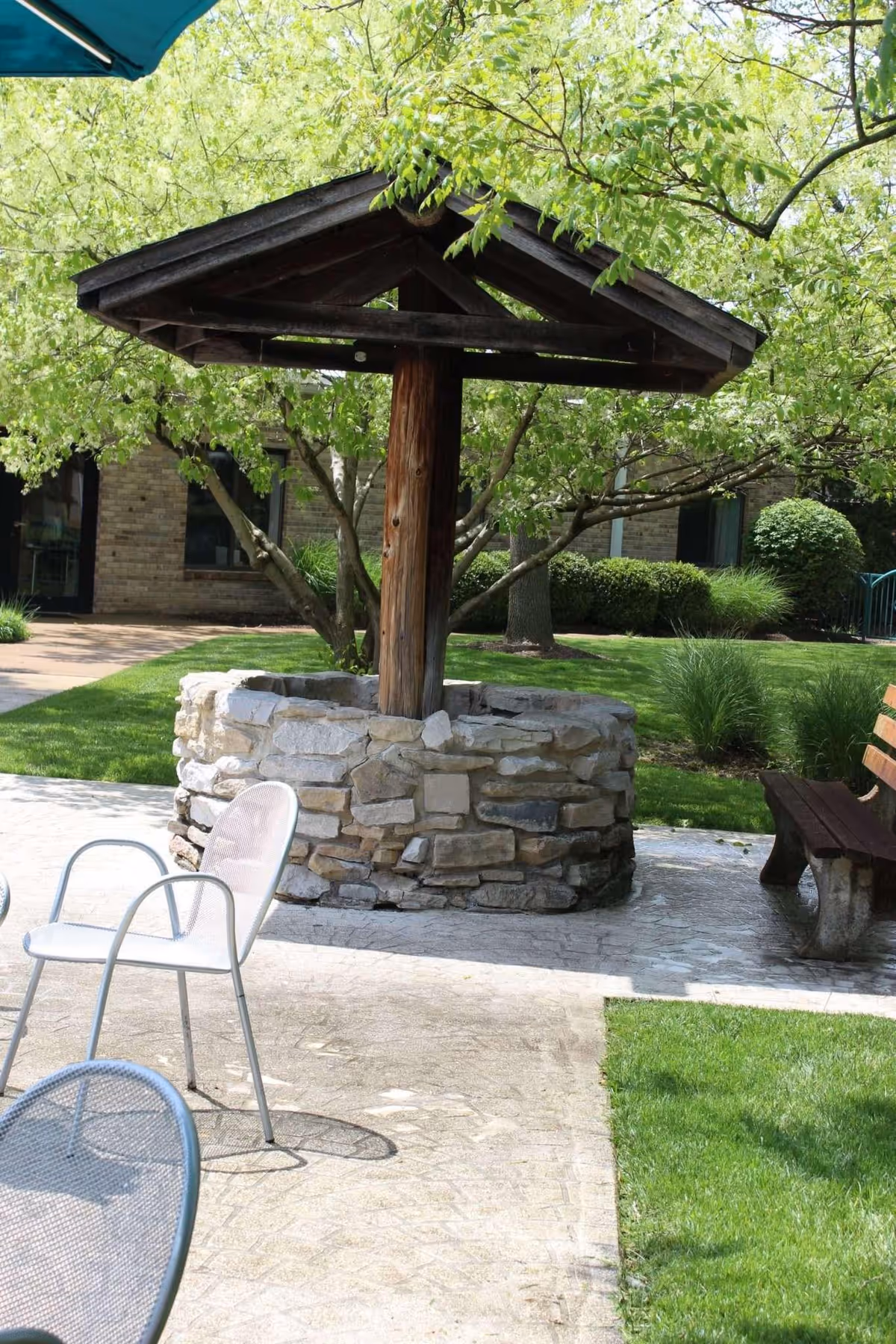 Outdoor seating area with metal chairs and a wooden bench near a stone well structure with a wooden roof, surrounded by green grass, trees, and shrubs in a garden setting.