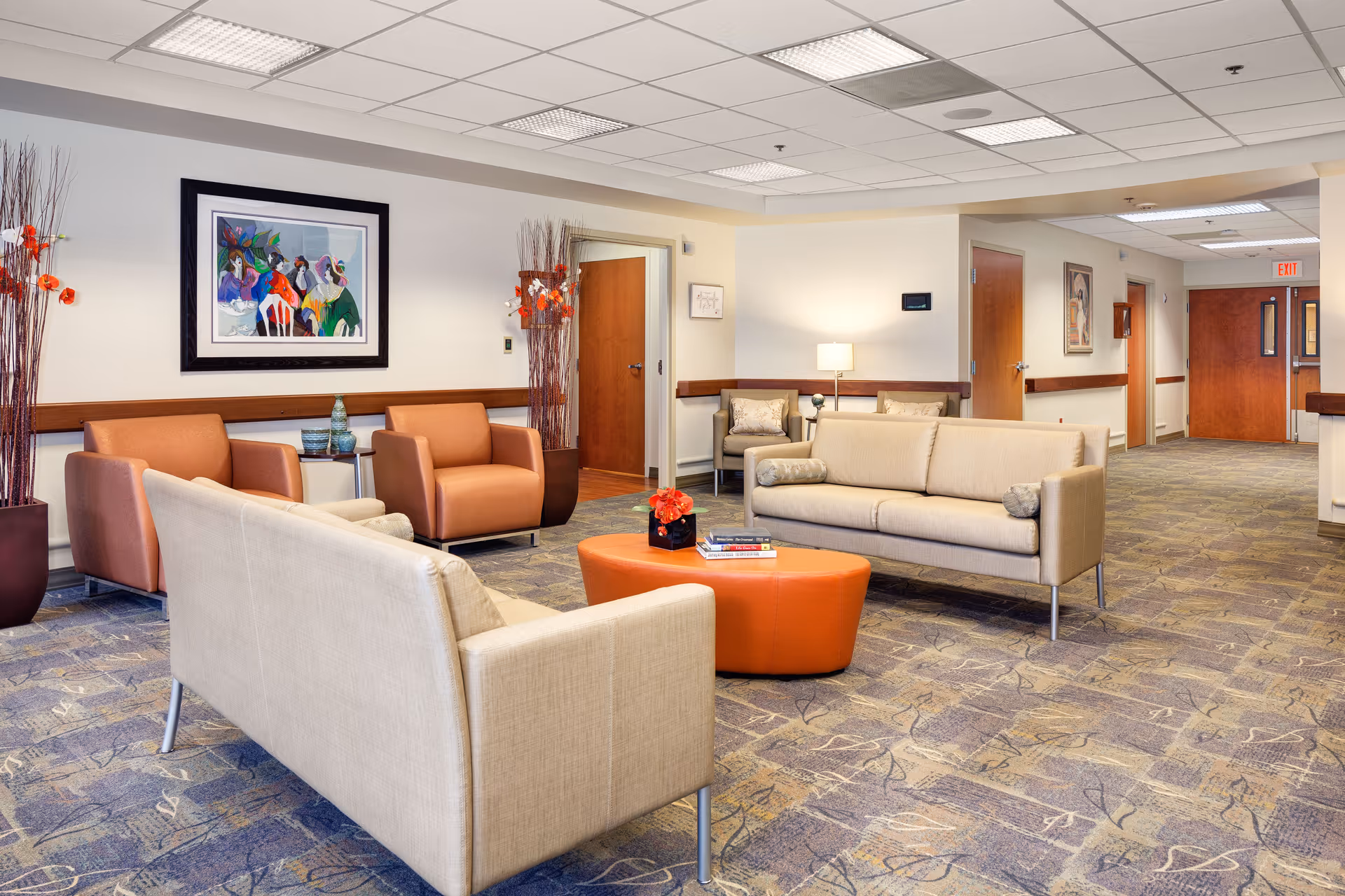 A spacious and well-lit common area with beige and orange armchairs and sofas arranged around an orange coffee table with books and a flower vase. The room has patterned carpet flooring, white walls with wooden trim, framed artwork, tall decorative plants, and several doors leading to other rooms. Ceiling lights illuminate the space.